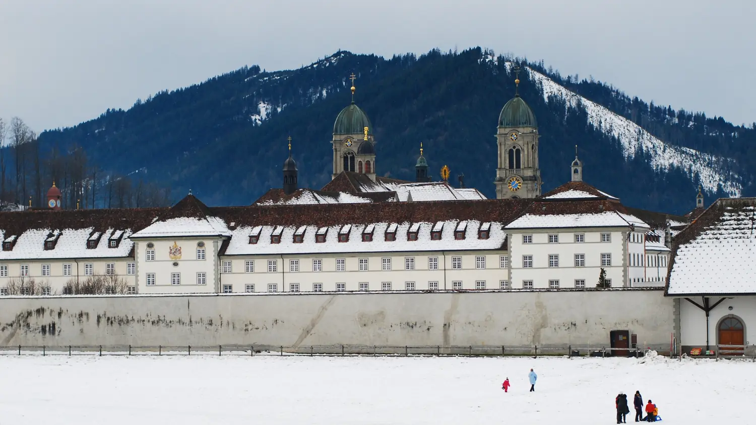 L'abbaye d'Einsiedeln sous la neige, devant elle des enfants avec des luges sur le champ enneigé