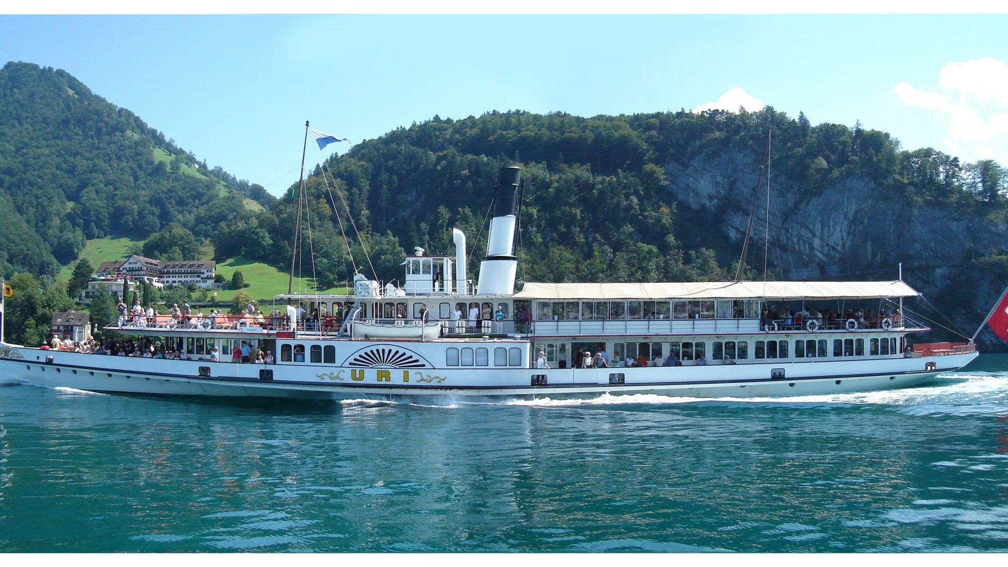 The paddle steamer Uri with a Swiss flag at the stern sails past a wooded shore on Lake Lucerne
